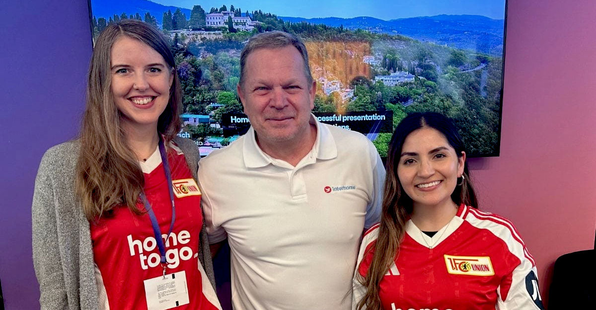 Drei Personen stehen lächelnd nebeneinander vor einem Bildschirm mit Landschaftsbild, zwei Frauen in roten „home to go“-Fußballtrikots und ein Mann in weißem Polohemd mit Interhome-Logo.
