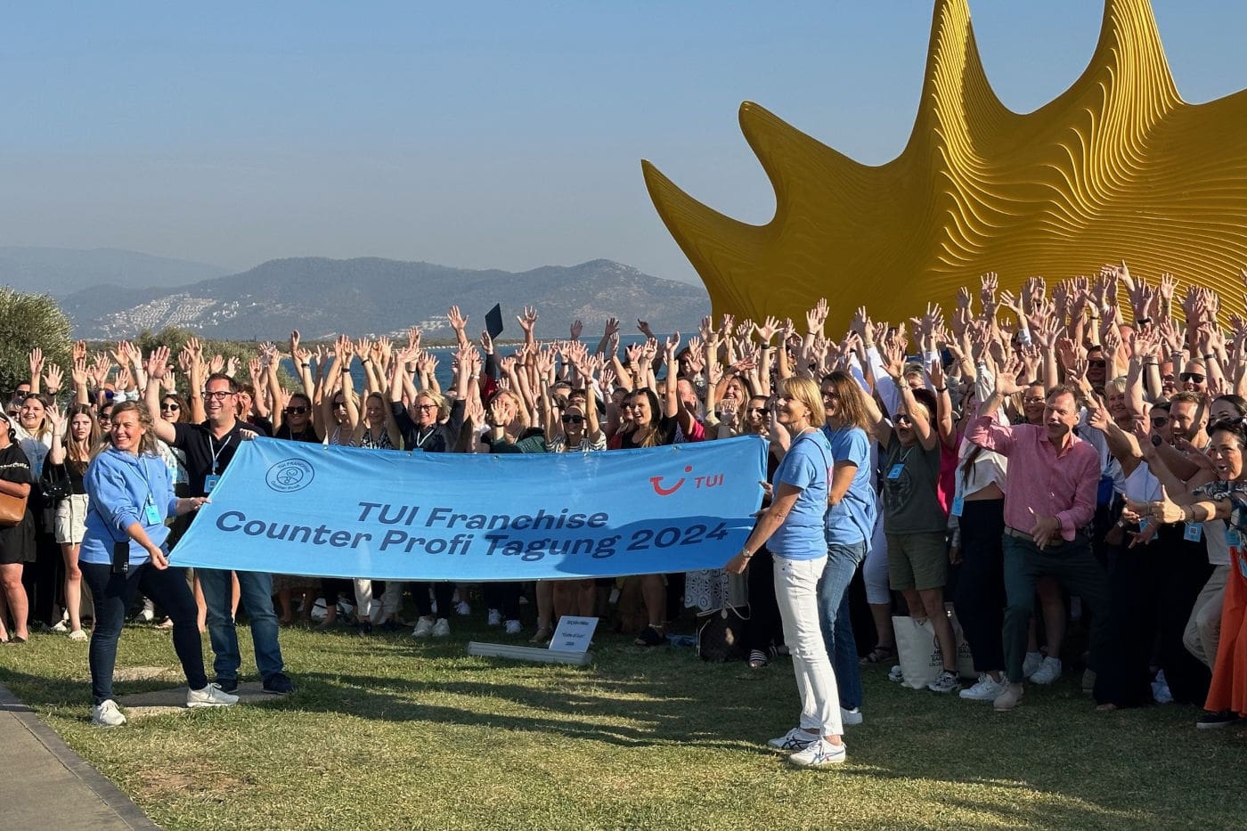Große Gruppe von Menschen im Freien, die gemeinsam ein blaues Banner mit der Aufschrift „TUI Franchise Counter Profi Tagung 2024“ halten. Viele Teilnehmende stehen dicht gedrängt dahinter und strecken jubelnd die Arme in die Luft. Im Hintergrund sind Berge, ein klarer Himmel und eine große, auffällige goldene Skulptur zu sehen.