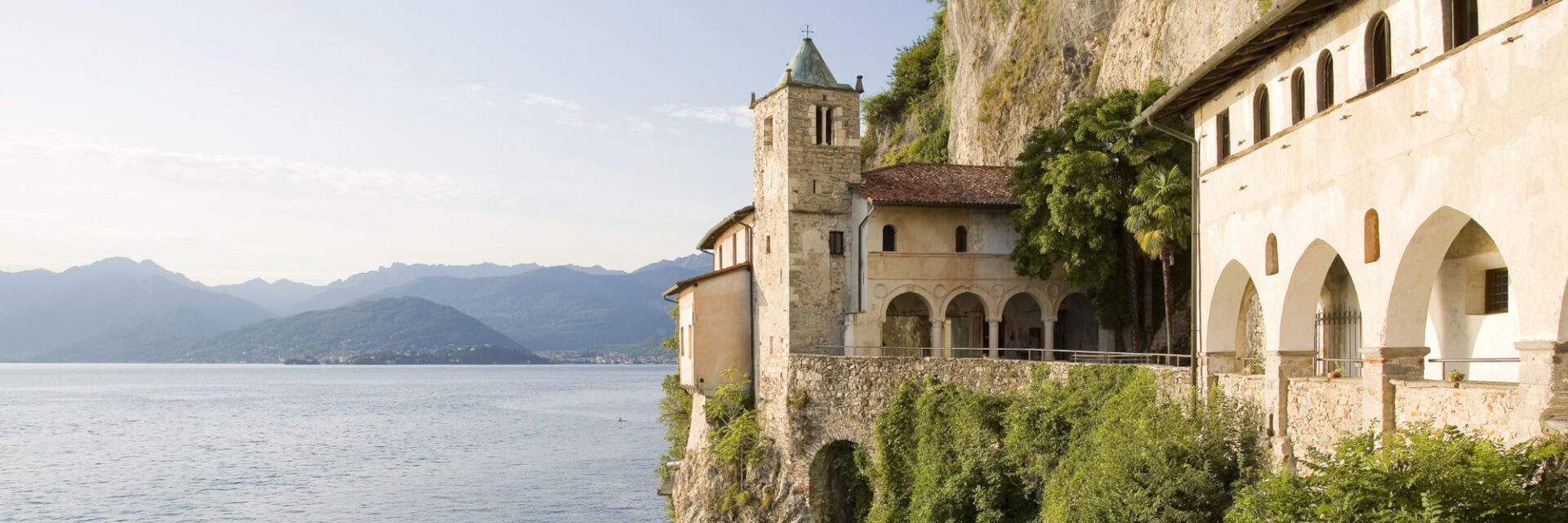 Eine Klosteranlage klebt an einer senkrechten Felswand über dem dunkelblauen Wasser des Lago Maggiore.