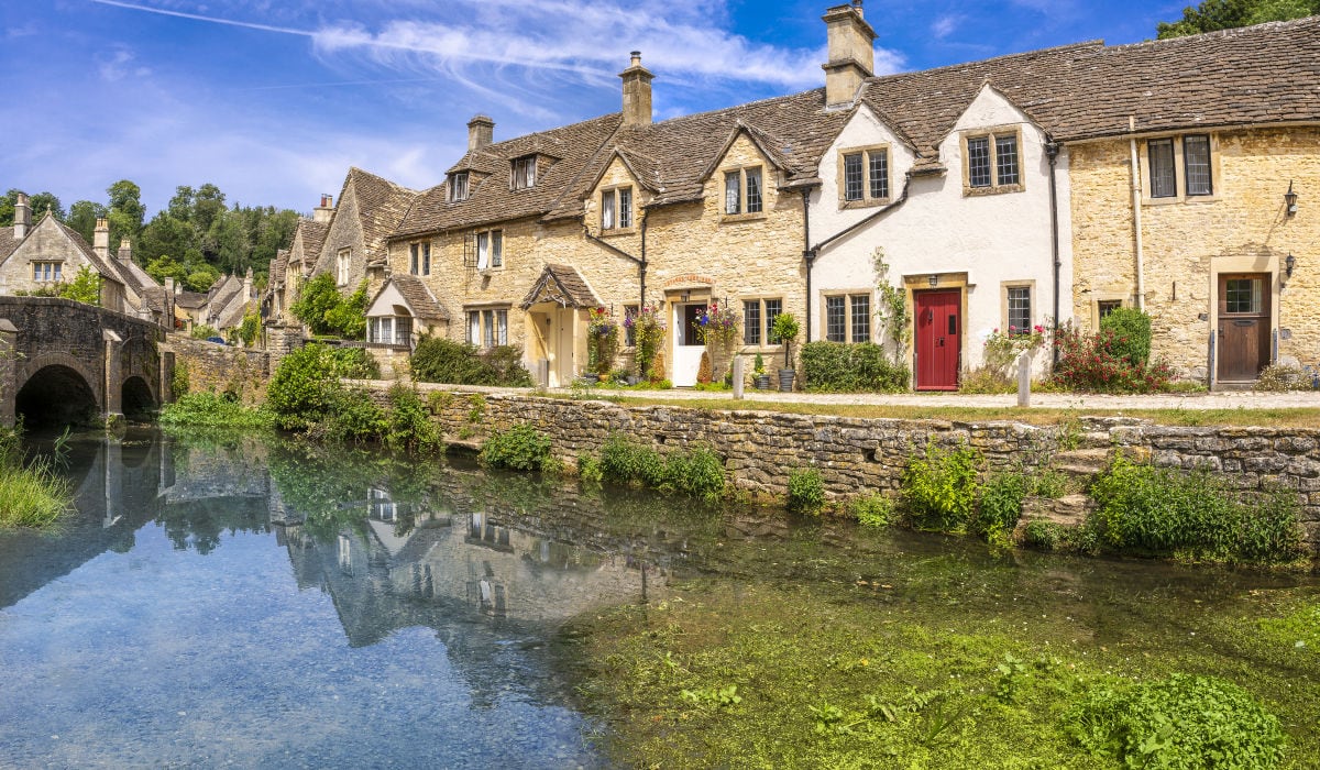 Idyllische Reihe historischer Natursteinhäuser in einem Dorf am Ufer eines klaren Baches, mit einer kleinen Steinbrücke, blühenden Pflanzen vor den Häusern und Spiegelungen der Gebäude im ruhigen Wasser unter blauem Himmel.