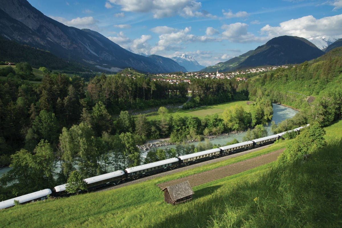 Ein langer Zug fährt durch ein grünes Tal mit Wiesen, Wald und einem Fluss, während im Hintergrund ein Dorf und hohe Berge unter einem blauen Himmel mit Wolken zu sehen sind.