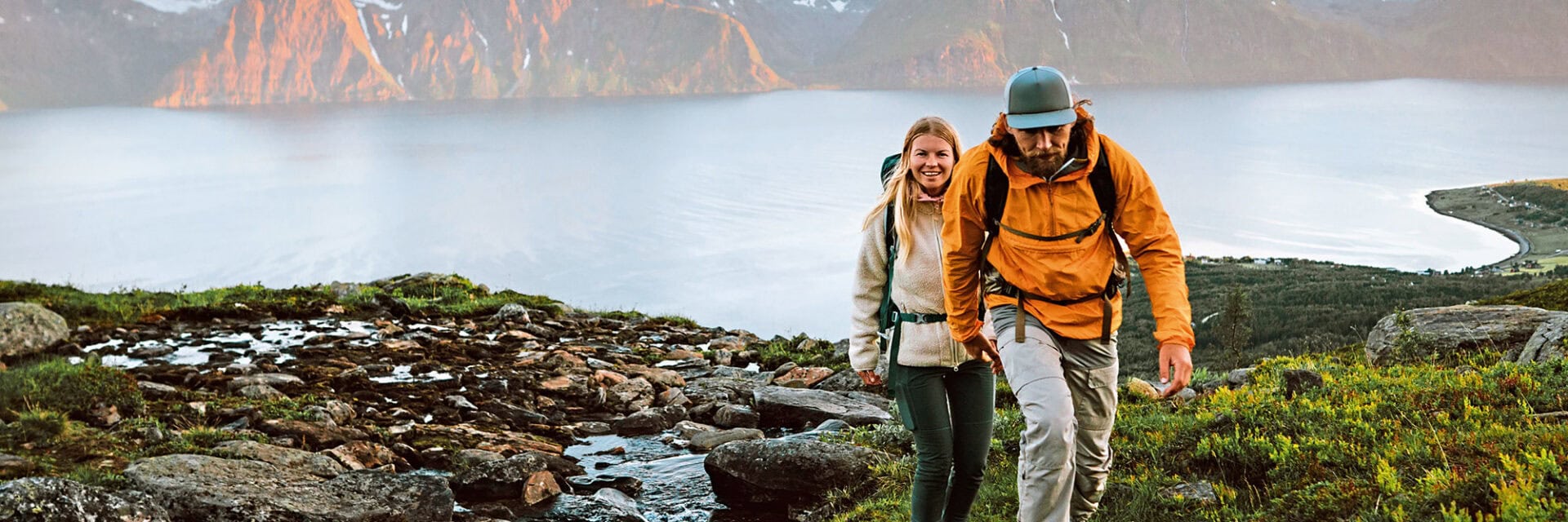 Ein Mann und eine Frau in Outdoor-Kleidung laufen neben einem Bach einen steilen Hang hinauf. Im Hintergrund sieht man einen großen See und steile Felswände in einem rötlichen Ton.