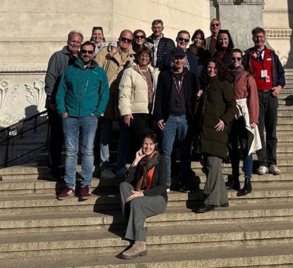 Gruppe von lächelnden Menschen steht auf den Stufen einer Treppe eines historischen Bauwerks im Sonnenschein.