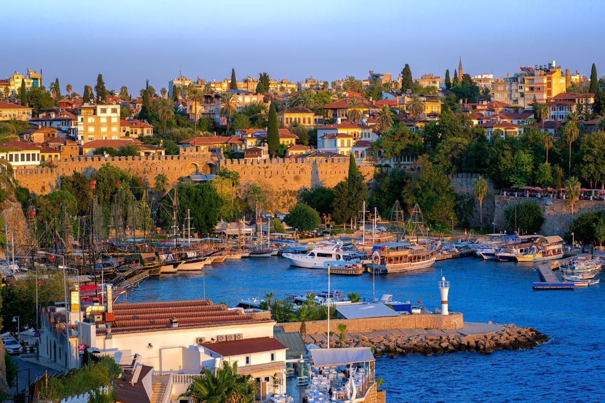 Blick auf einen Hafen bei warmem Abendlicht: Im Vordergrund liegen mehrere Ausflugsboote und Yachten im blauen Wasser. Dahinter erhebt sich eine historische Stadtmauer aus Stein, über der dicht bebaute Häuser mit roten Ziegeldächern, Palmen und hohe Bäume zu sehen sind. Die Szene wirkt mediterran und sonnig.