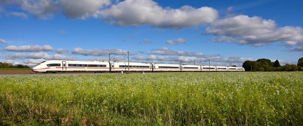 Ein moderner ICE-Schnellzug fährt bei Tageslicht durch eine grüne, weite Landschaft unter blauem Himmel mit weißen Wolken. Das Bild zeigt Zugreise, Bahnverkehr und Natur in Deutschland.
