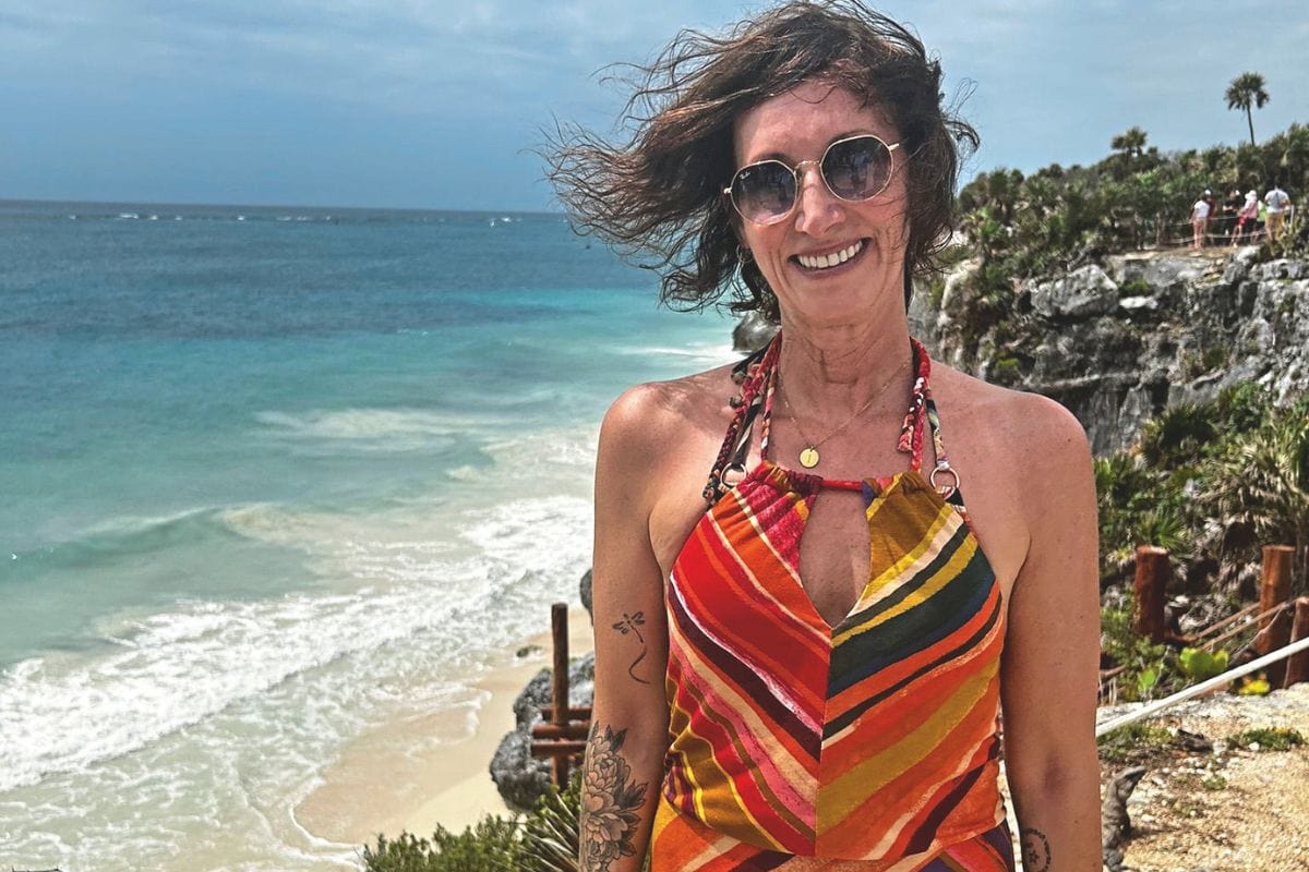 Eine lächelnde Frau mit Sonnenbrille und buntem Kleid steht vor einer felsigen Küste mit blauem Meer und weißem Sandstrand im Hintergrund bei sonnigem Wetter.