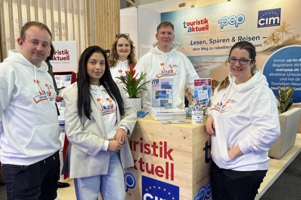 Fünf junge Erwachsene in weißen „Strandtherapeut“-Hoodies stehen an einem Messestand von „touristik aktuell“ und „meinpep“ auf einer Tourismusmesse. Auf dem Tresen liegen Magazine und Flyer, im Hintergrund sind Werbebanner mit Strandmotiv zu sehen.