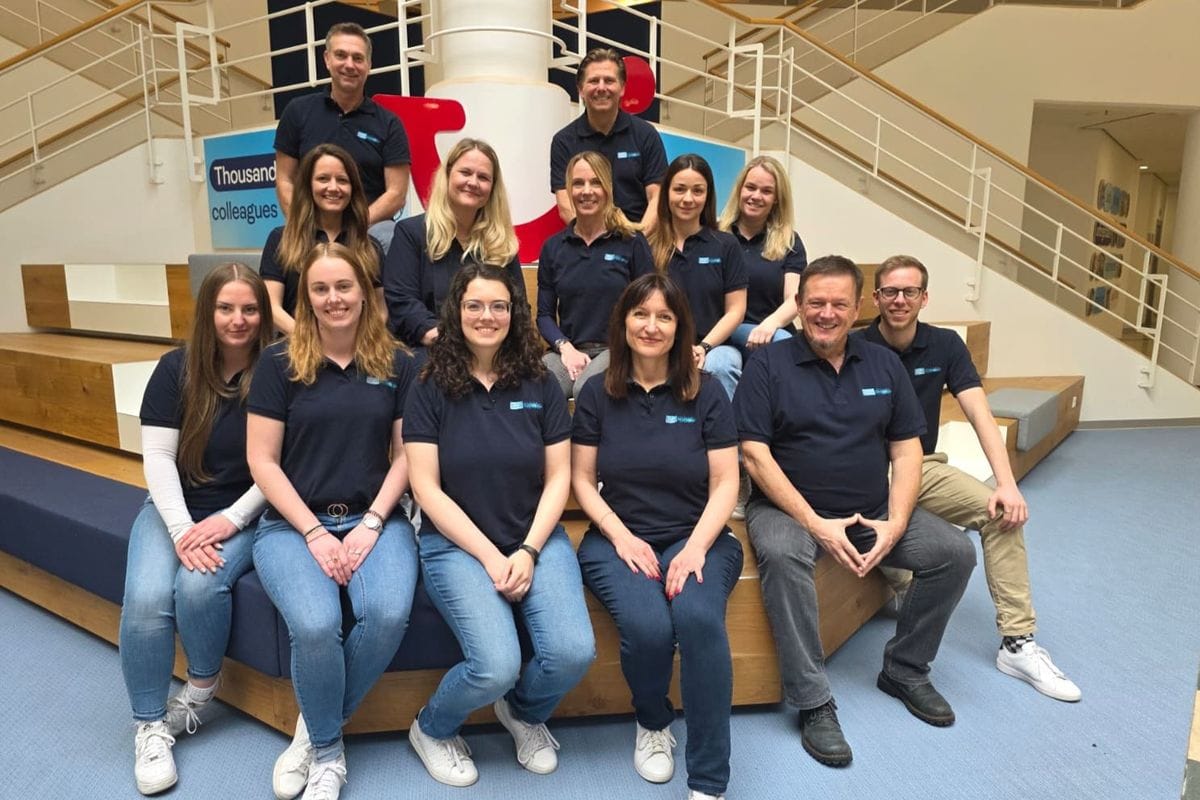 Gruppe von Mitarbeitenden in dunkelblauen TUI-Campus-Polohemden sitzt und steht auf Holzstufen in einem modernen, hellen Atrium mit Treppen und Firmenbeschilderung im Hintergrund.