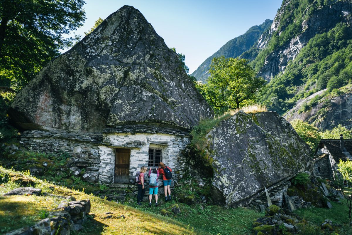 Wanderer vor traditionellen Steinhäusern unter großen Felsblöcken im Val Calnègia im Tessin, umgeben von alpiner Berglandschaft.