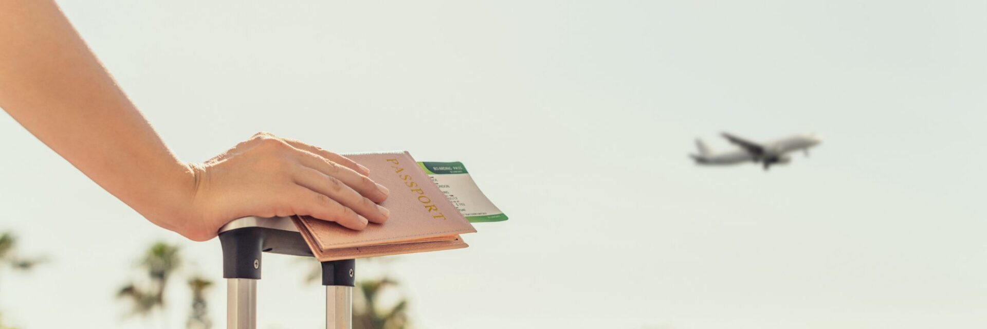 Großaufnahme einer Hand mit Passport und Flugticket in der Hand, die sich aufn einem Koffer abstützt. Im Hintergrund fliegt ein Flugzeug.