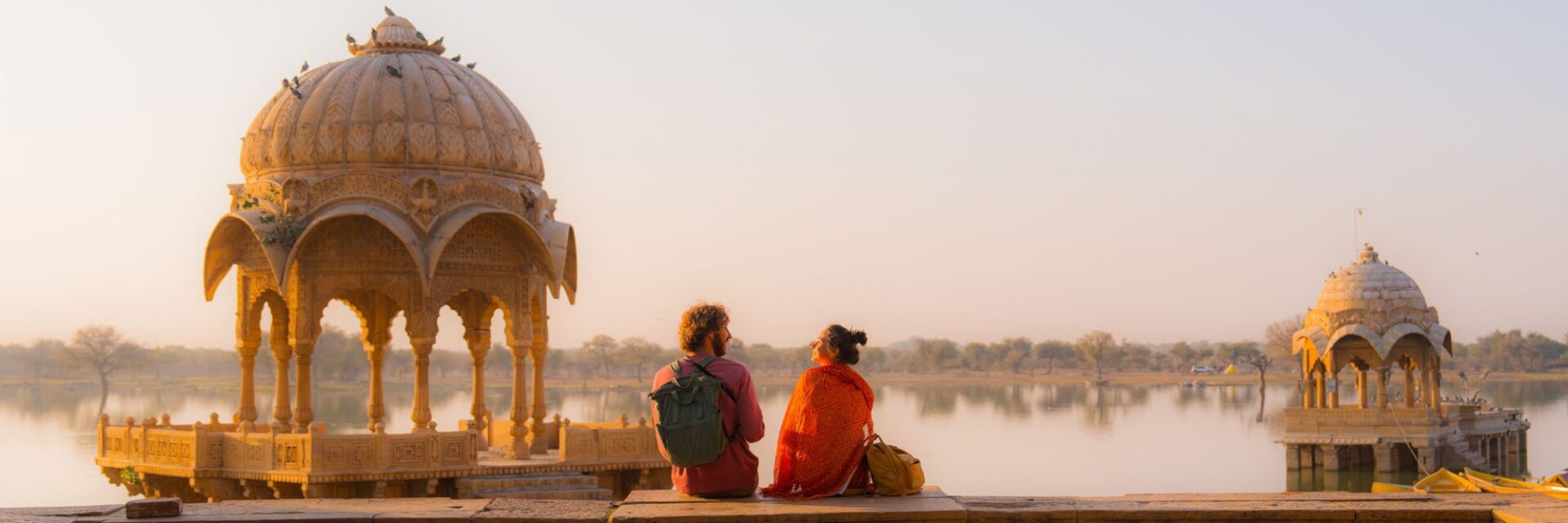 Zwei Touristen sitzen am Ufer eines Sees in Indien vor kunstvollen Sandsteinpavillons.