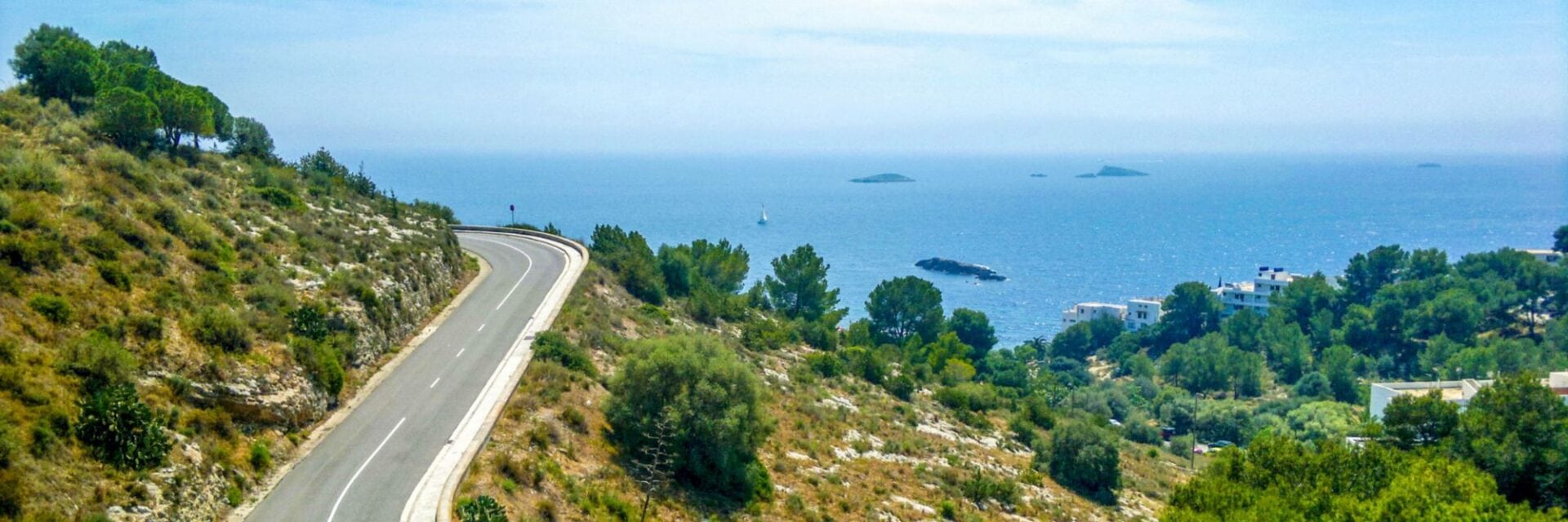 Geschwungene Straße entlang einer grünen Hügellandschaft auf Ibiza mit Blick auf das Meer und kleine Inseln im Hintergrund.