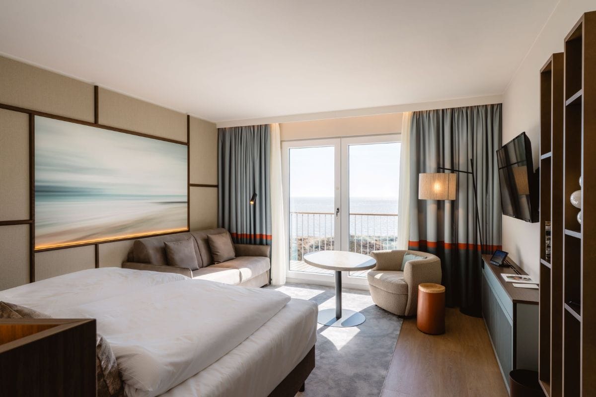 Modernes Hotelzimmer auf Sylt mit Meerblick: Das Bild zeigt ein helles, stilvoll eingerichtetes Doppelzimmer mit großem Bett, gemütlichem Sofa, Sessel, Flachbildfernseher und bodentiefem Fenster, das den Blick auf die Nordsee freigibt.