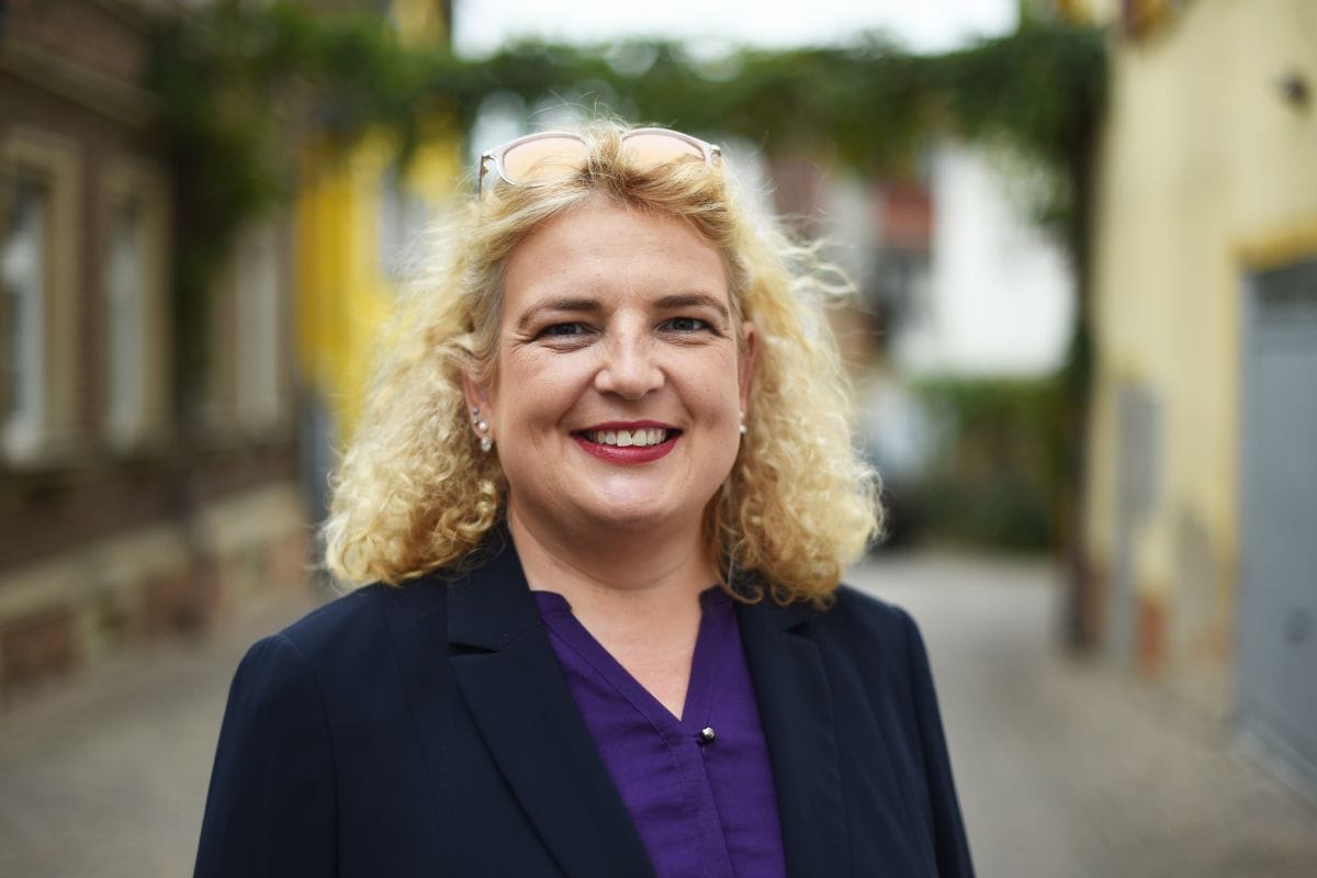 Porträt einer lächelnden Frau mit lockigem blondem Haar, dunklem Blazer und lila Bluse, aufgenommen in einer ruhigen Straße mit unscharfem Hintergrund.