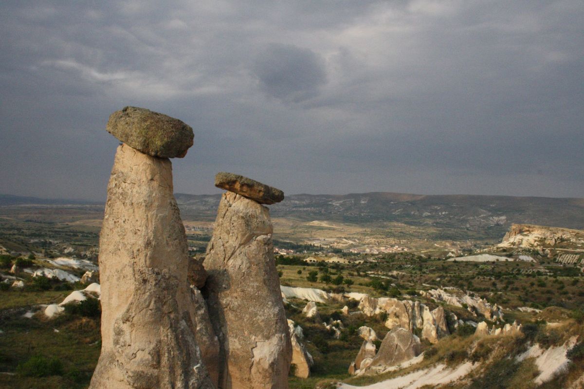 Zwei markante Feenkamine mit großen Felsbrocken auf den Spitzen ragen in die Höhe, dahinter breitet sich die beeindruckende Landschaft Kappadokiens in der Türkei aus. Der Himmel ist bewölkt und verleiht der Szene eine mystische Stimmung. Das Bild zeigt die einzigartigen Felsformationen, eine beliebte Sehenswürdigkeit für Natur- und Wanderurlaub in der Türkei.