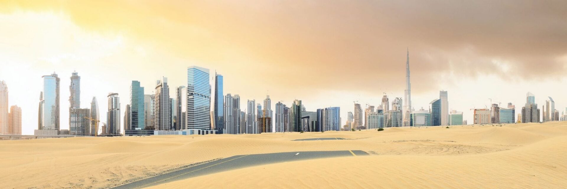 buchungsdelle Weite Wüstenlandschaft mit gelbem Sand im Vordergrund, dahinter die moderne Skyline von Dubai mit vielen Hochhäusern und einem sehr hohen Turm unter dramatisch bewölktem Himmel bei Sonnenuntergang.