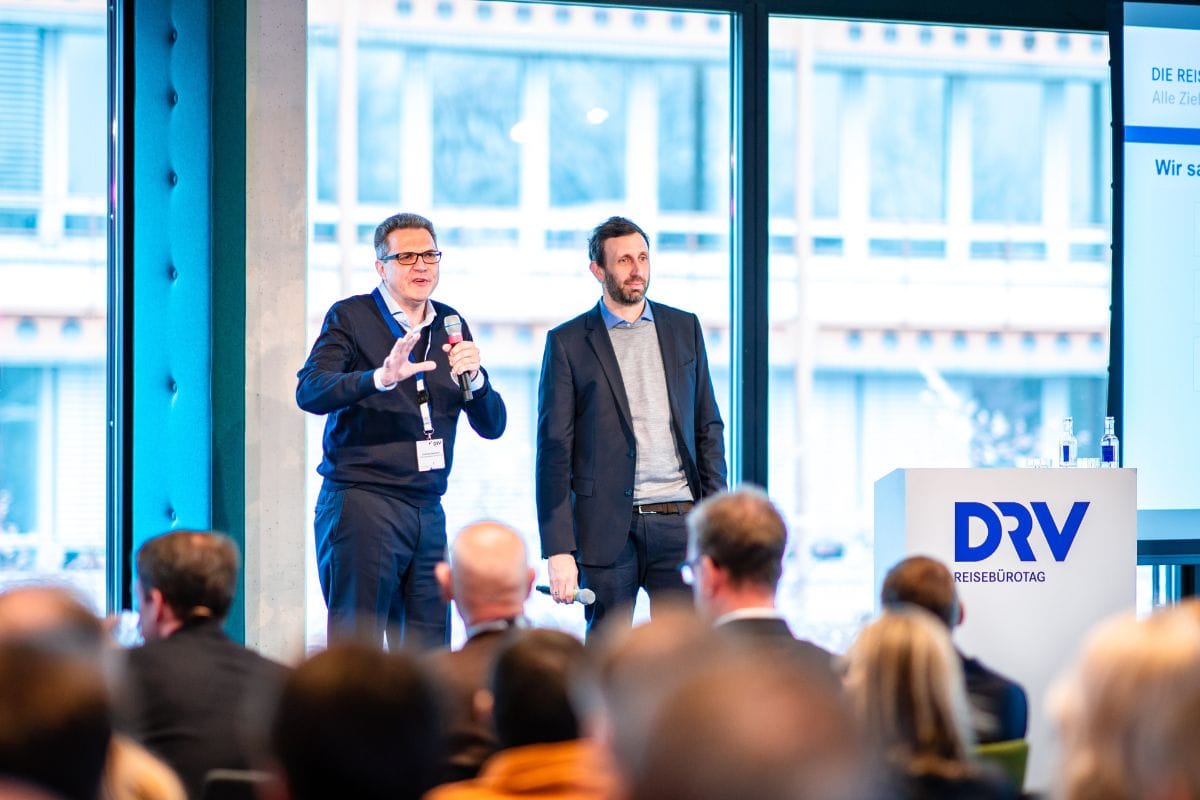 Zwei Männer stehen auf einer Bühne vor sitzenden Menschen und moderieren