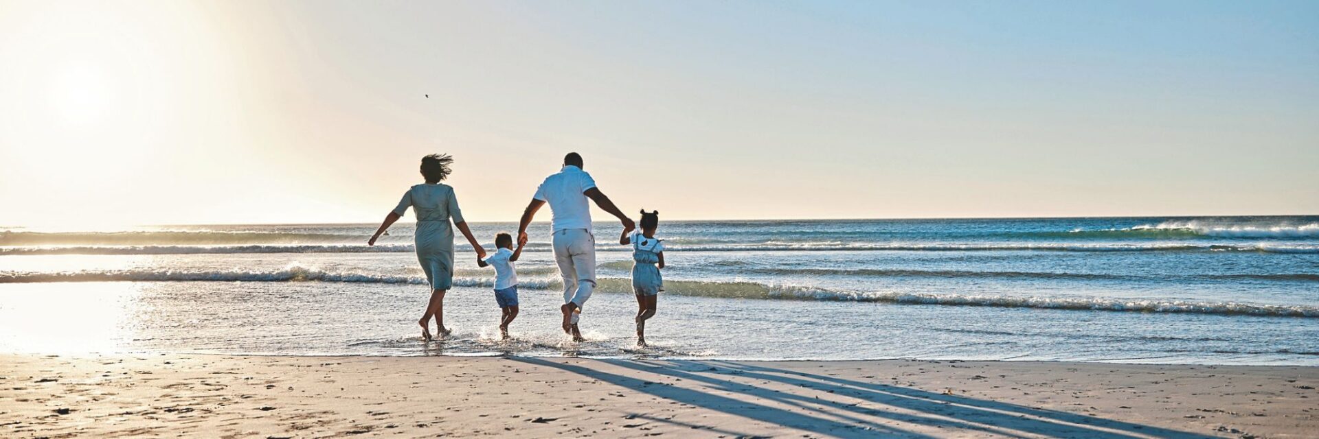 Eine vierköpfige Familie läuft Hand in Hand am Strand ins flache Meer, die Sonne steht tief am Himmel und wirft lange Schatten auf den Sand, während sanfte Wellen ans Ufer rollen.