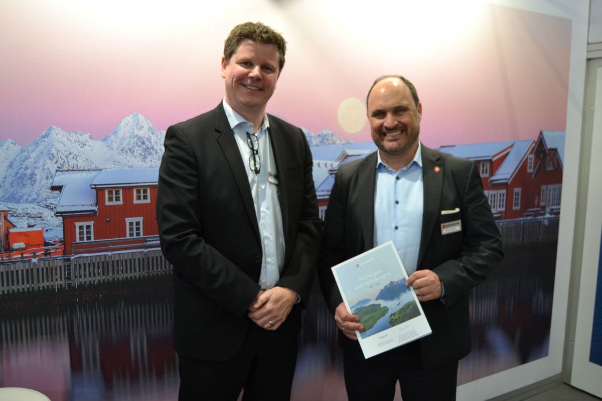 Zwei Reederei-Manager mittleren Alters präsentieren auf einem Messestand einen neuen Reisekatalog. Sie blicken lächelnd in die Kamera.