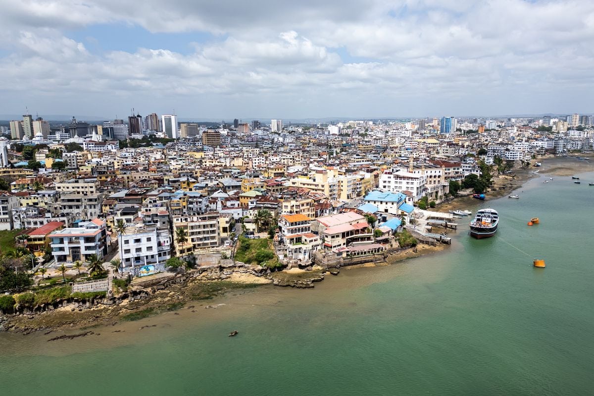 Luftaufnahme der Küstenstadt Mombasa mit dicht bebauten Häusern, Hochhäusern im Hintergrund und einem Boot vor der Uferlinie im türkisfarbenen Indischen Ozean.
