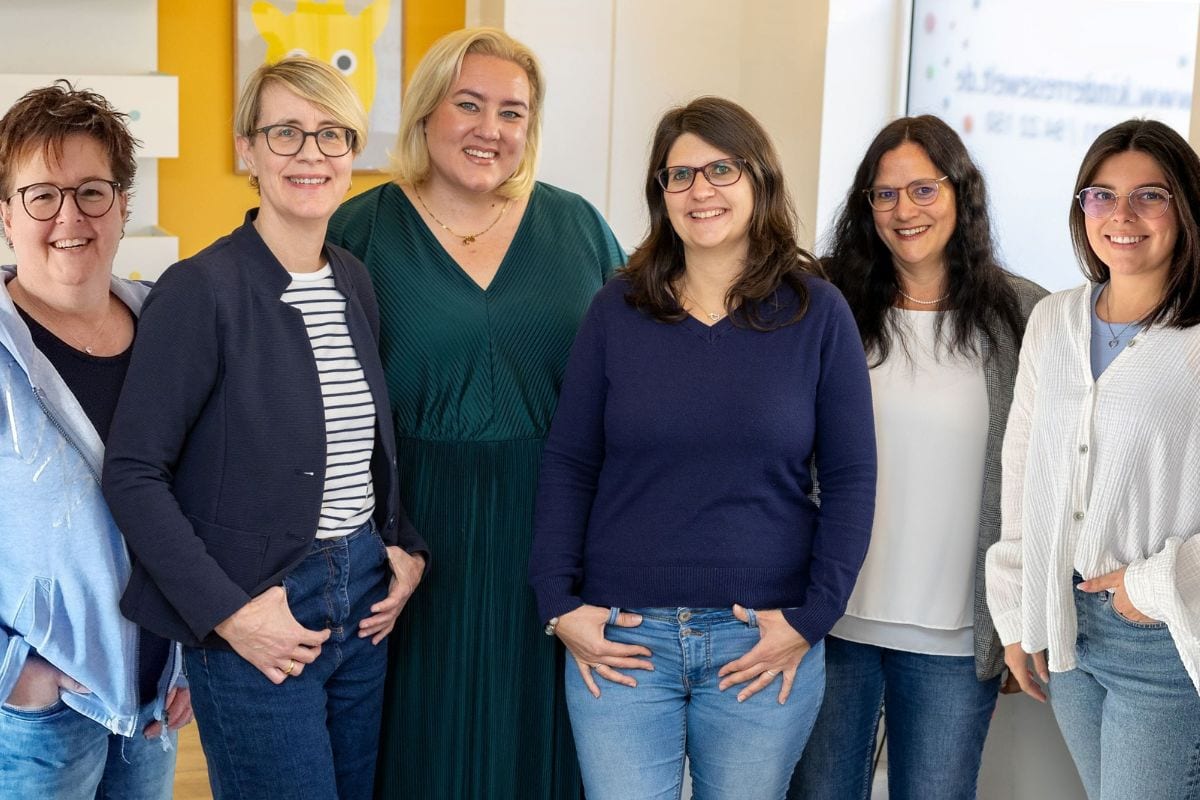 Sechs Frauen stehen nebeneinander in einem modernen, hellen Raum, einige tragen Jeans. Im Hintergrund sind bunte Wanddekorationen und ein Bildschirm mit Schrift zu sehen.
