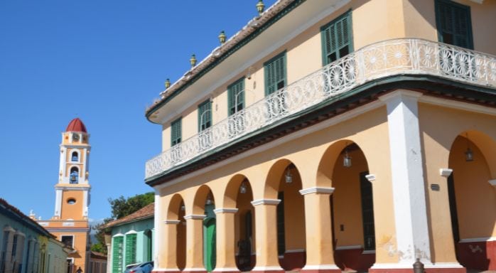 Historisches Gebäude im Kolonialstil in Trinidad, Kuba mit gelb gestrichener Fassade und grünen Fensterläden, daneben der markante Glockenturm der Iglesia de San Francisco. Das Bild zeigt typische Architektur von Trinidad, einer Unesco-Weltkulturerbestadt in Kuba, bei strahlend blauem Himmel.