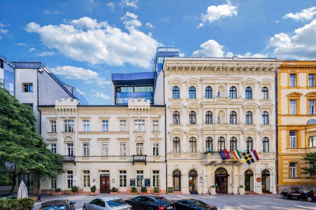 Das Bild zeigt die historische Fassade des Melia Hotels in Budapest bei Tageslicht, mit mehreren wehenden Länderflaggen über dem Eingangsbereich. Vor dem Gebäude parken Autos und der Himmel ist blau mit dekorativen Wolken.