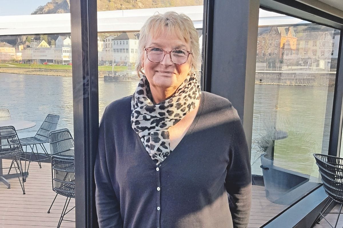 Eine Frau mit blonden kurzen Haaren, einer Brille und einem Schal im Leoparden-Look steht in einem Raum auf einem Flusskreuzfahrtschiff und lächtelt in die Kamera