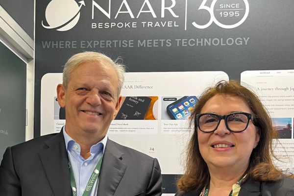 Zwei Personen stehen lächelnd vor einem Messestand mit dem Logo „Naar Bespoke Travel“. Im Hintergrund sind ein Bildschirm und Informationsgrafiken zu sehen, darüber der Slogan „Where Expertise Meets Technology“. Beide tragen Businesskleidung und Messeausweise.