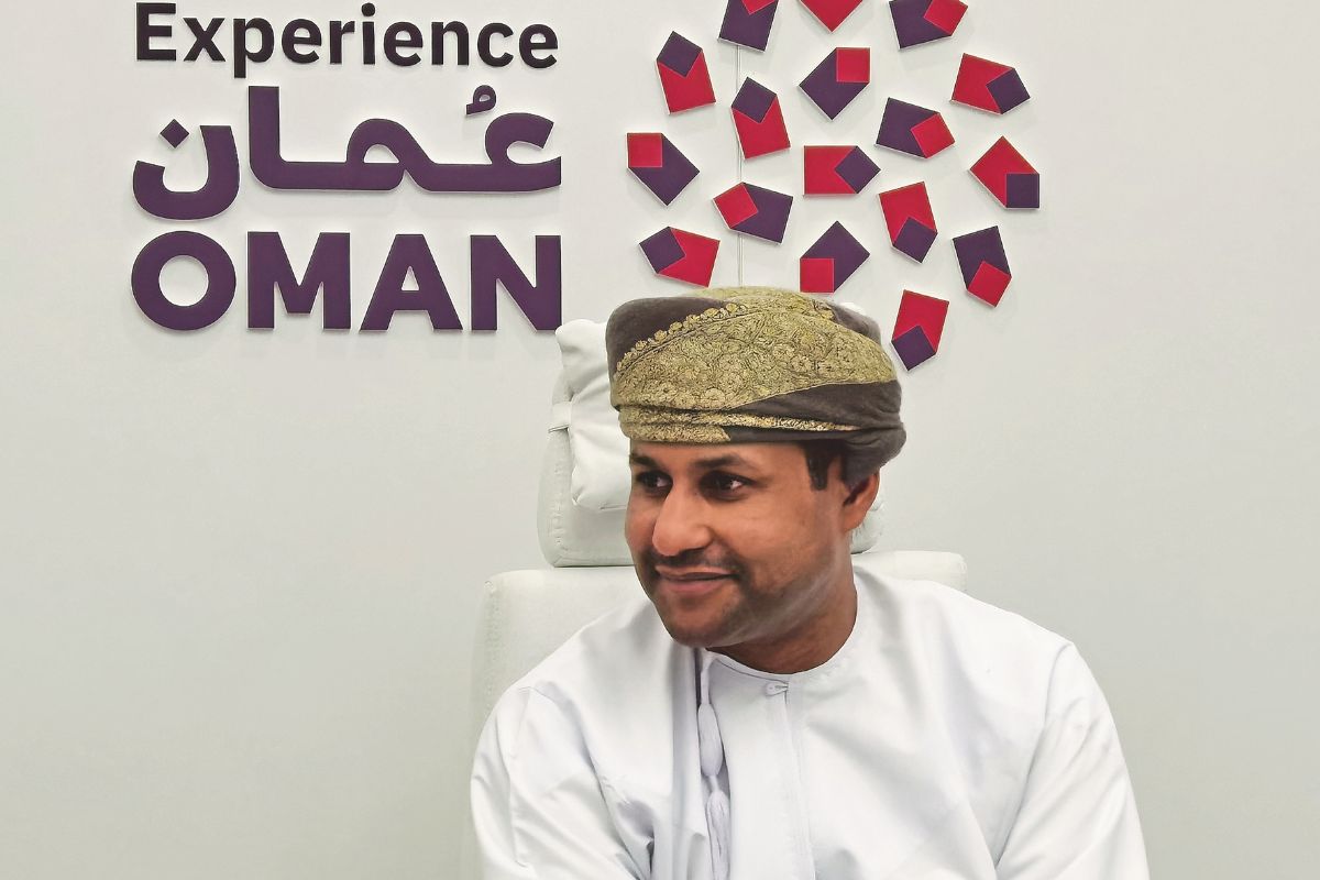 Ein Mann in traditioneller omanischer Kleidung sitzt vor einem Laptop in einem modernen Messestand; im Hintergrund steht der Schriftzug „Experience Oman“ auf Arabisch und Englisch sowie eine dekorative Wandgestaltung in Rot und Violett.