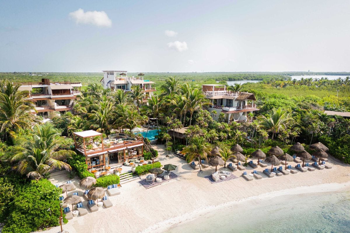 Das Foto zeigt ein luxuriöses Strandhotel in Tulum, Mexiko, umgeben von tropischen Palmen und direkt am weißen Sandstrand mit Sonnenliegen und Palapa-Sonnenschirmen. Im Hintergrund ist dichter Dschungel zu sehen, während das türkisfarbene Meer sanft ans Ufer trifft.