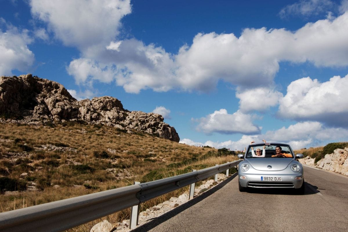 Zwei Menschen fahren lachend in einem silbernen Cabrio entlang einer einsamen Landstraße durch eine felsige Hügellandschaft unter blauem Himmel mit großen weißen Wolken.