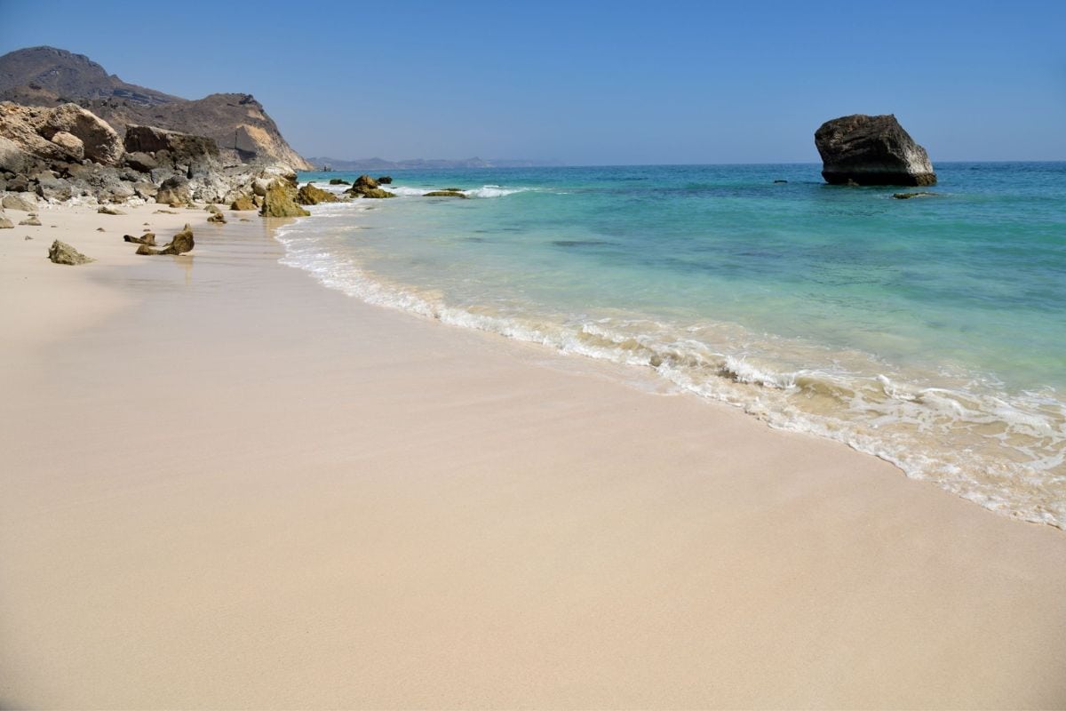 Weißer Sandstrand mit sanften Wellen am türkisfarbenen Meer, im Hintergrund felsige Klippen und ein einzelner großer Felsen im Wasser, klare Sicht und strahlend blauer Himmel