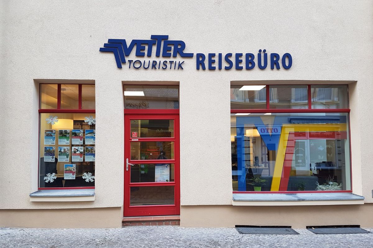 Außenansicht eines Reisebüros mit der Beschriftung „Vetter Touristik Reisebüro“, roter Eingangstür und Schaufenstern mit Reiseprospekten.