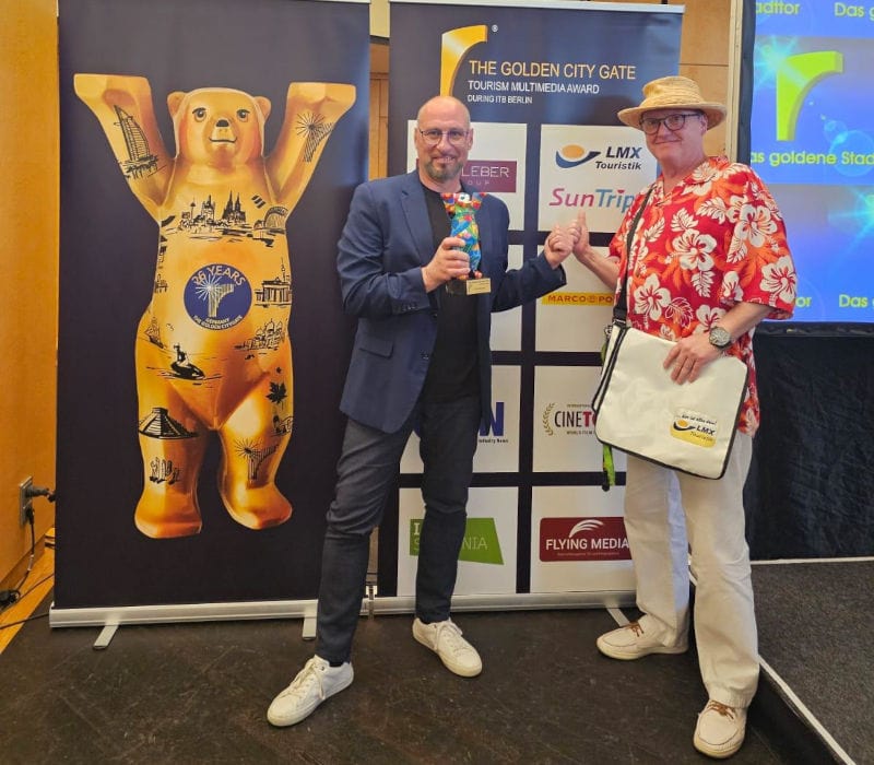 Zwei Männer stehen vor einem Veranstaltungsbanner und posieren für ein Foto. Der Mann links trägt ein dunkles Sakko, Jeans und weiße Sneaker und hält eine bunte Trophäe in der Hand. Der Mann rechts trägt ein rotes Hawaiihemd, einen Strohhut, helle Hose und eine Umhängetasche und zeigt mit dem Finger auf die Auszeichnung. Neben ihnen ist eine große goldene Bärenfigur mit erhobenen Armen zu sehen. Im Hintergrund sind Logos und der Schriftzug „The Golden City Gate – Tourism Multimedia Award“ erkennbar.