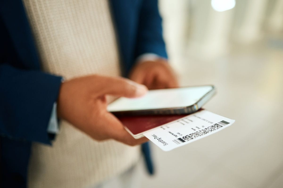 Eine Person hält am Flughafen ein Flugticket, einen Reisepass und ein Smartphone in der Hand, bereit zum Boarding. Das Foto illustriert das Thema Flugreise, Reisedokumente und digitale Bordkarten. Im Vordergrund stehen die wichtigsten Utensilien für internationale Reisen.
