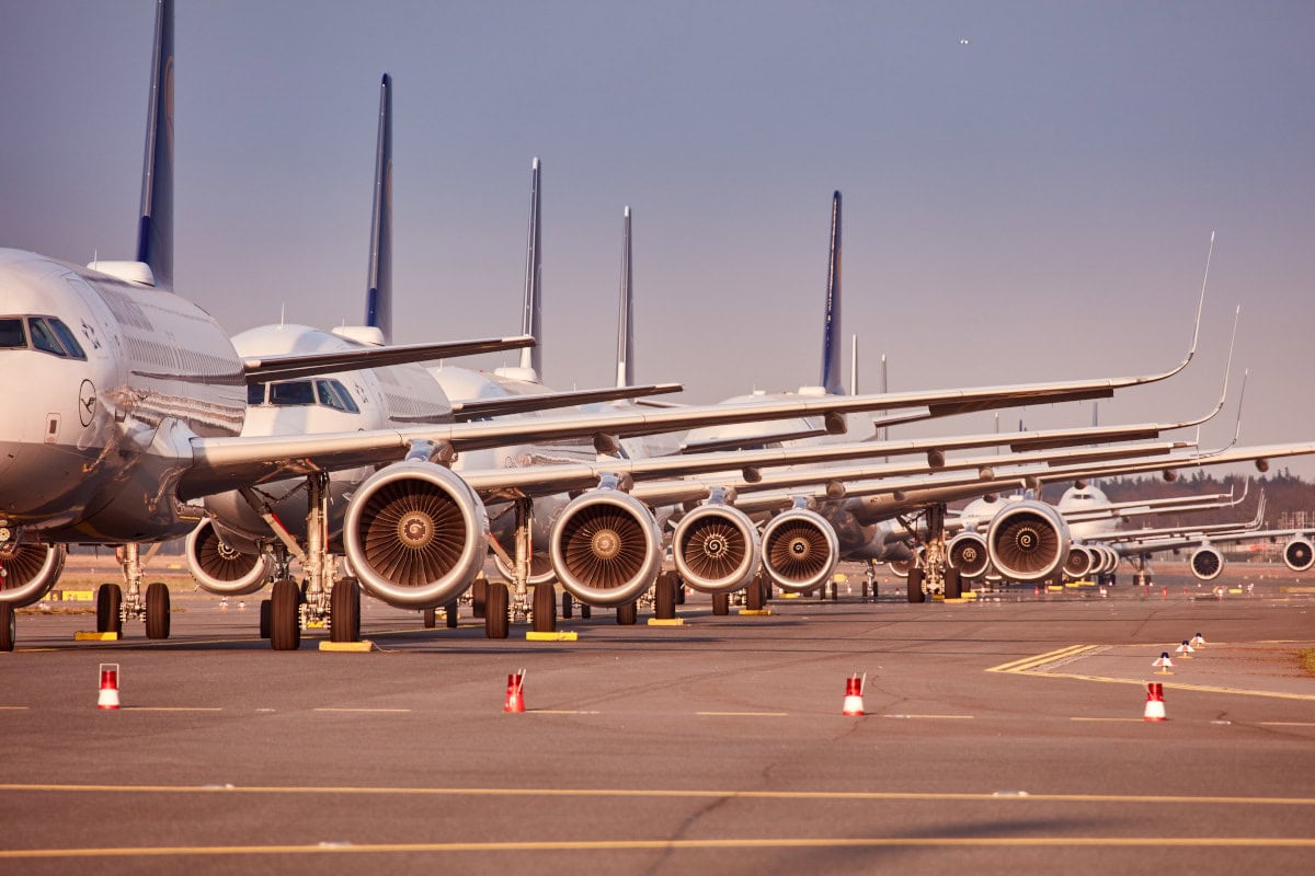 Mehrere Lufthansa-Flugzeuge stehen dicht nebeneinander auf einem Flughafen-Parkplatz, die Triebwerke und die Flugzeugnasen sind prominent sichtbar. Das Bild zeigt die parkenden Jets im Sonnenlicht, während die Start- und Landebahn im Hintergrund zu erkennen ist.