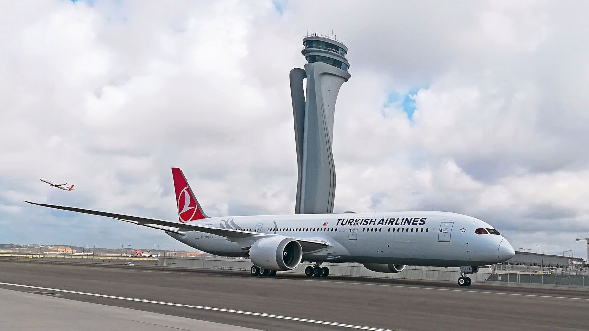 Ein Turkish-Airlines-Flugzeug vom Typ Dreamliner rollt oder steht auf dem Vorfeld vor dem markanten Kontrollturm des Flughafens; im Hintergrund hebt ein weiteres Flugzeug ab.