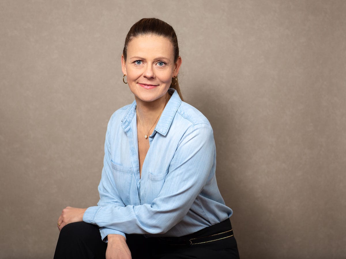 Auf dem Foto sitzt eine Frau in geschäftlicher Kleidung, bestehend aus einer hellblauen Bluse und einer schwarzen Hose, vor einem neutralen hellbraunen Hintergrund. Sie hat ihre Haare streng nach hinten gebunden und trägt schlichte Schmuckstücke wie eine Halskette und Ohrringe.