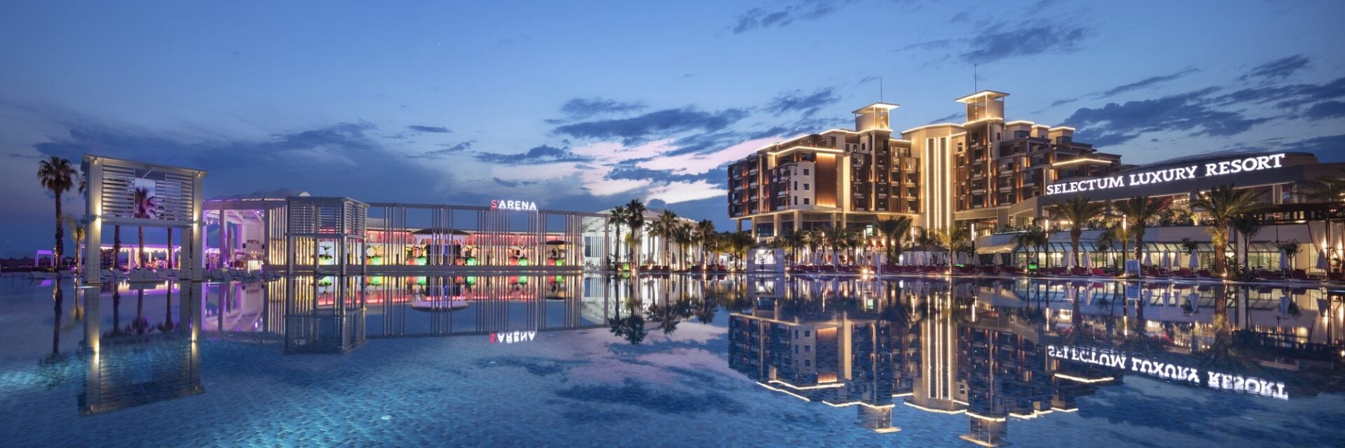 Blick über einen Pool auf eine sehr große Hotelanlage im Abendlicht