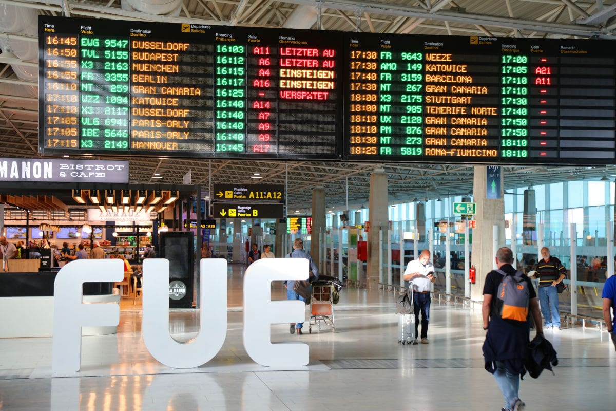 Im modernen Terminal des Flughafens Fuerteventura stehen große weiße Buchstaben mit der Abkürzung „FUE“ vor einer lebhaften Abflugtafel, die zahlreiche Flüge nach europäischen Zielen wie Düsseldorf, Budapest und Barcelona anzeigt. Im Hintergrund bewegen sich Reisende durch die helle, offene Halle, während das Bistro und Café und verschiedene Wegweiser für Orientierung sorgen.