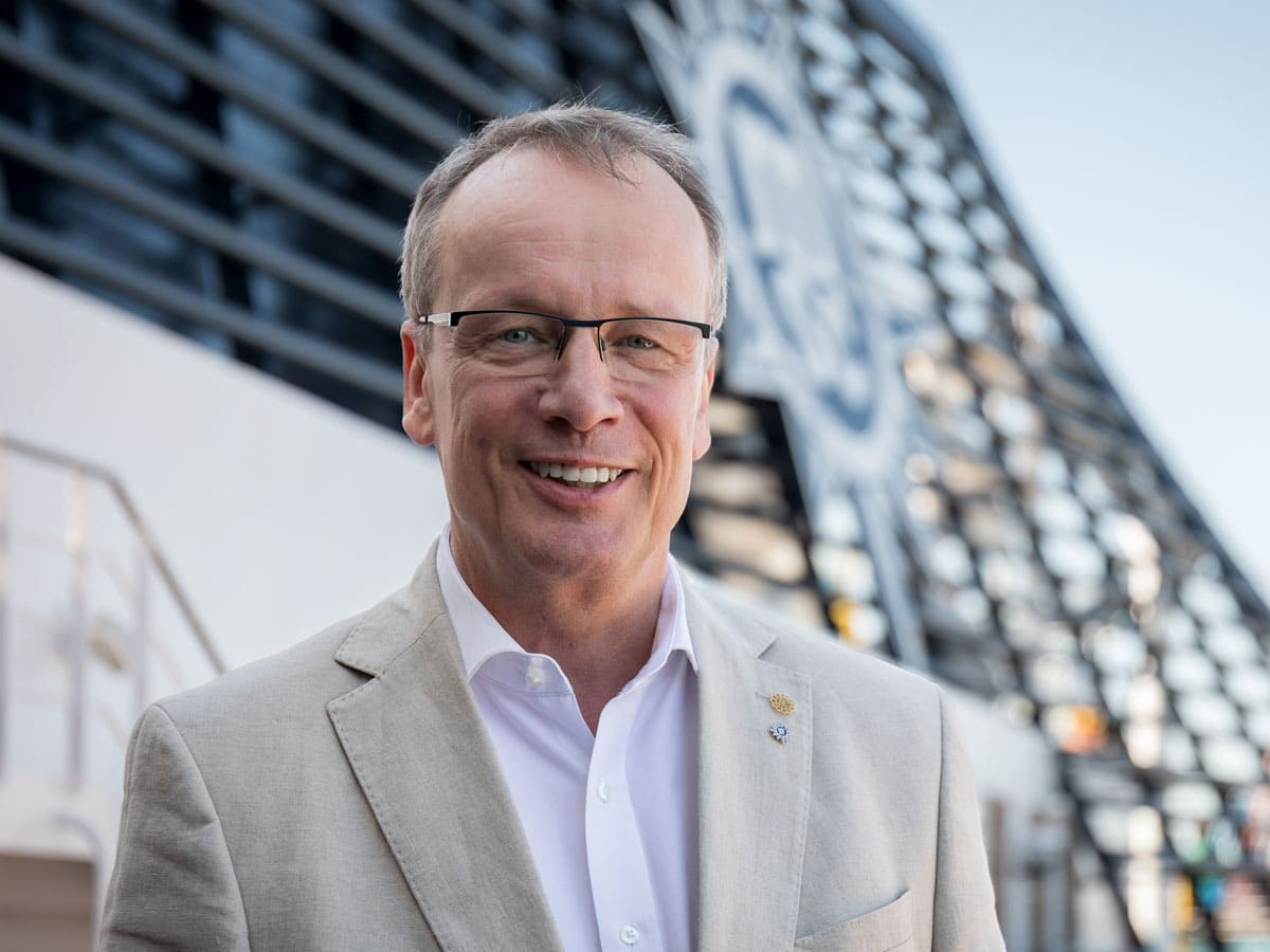 Ein Reederei-Manager mit Brille und hellem Sakko steht lächelnd auf einem Kreuzfahrtschiff. Im Hintergrund ist das blaue Logo der Reederei MSC zu erkennen.