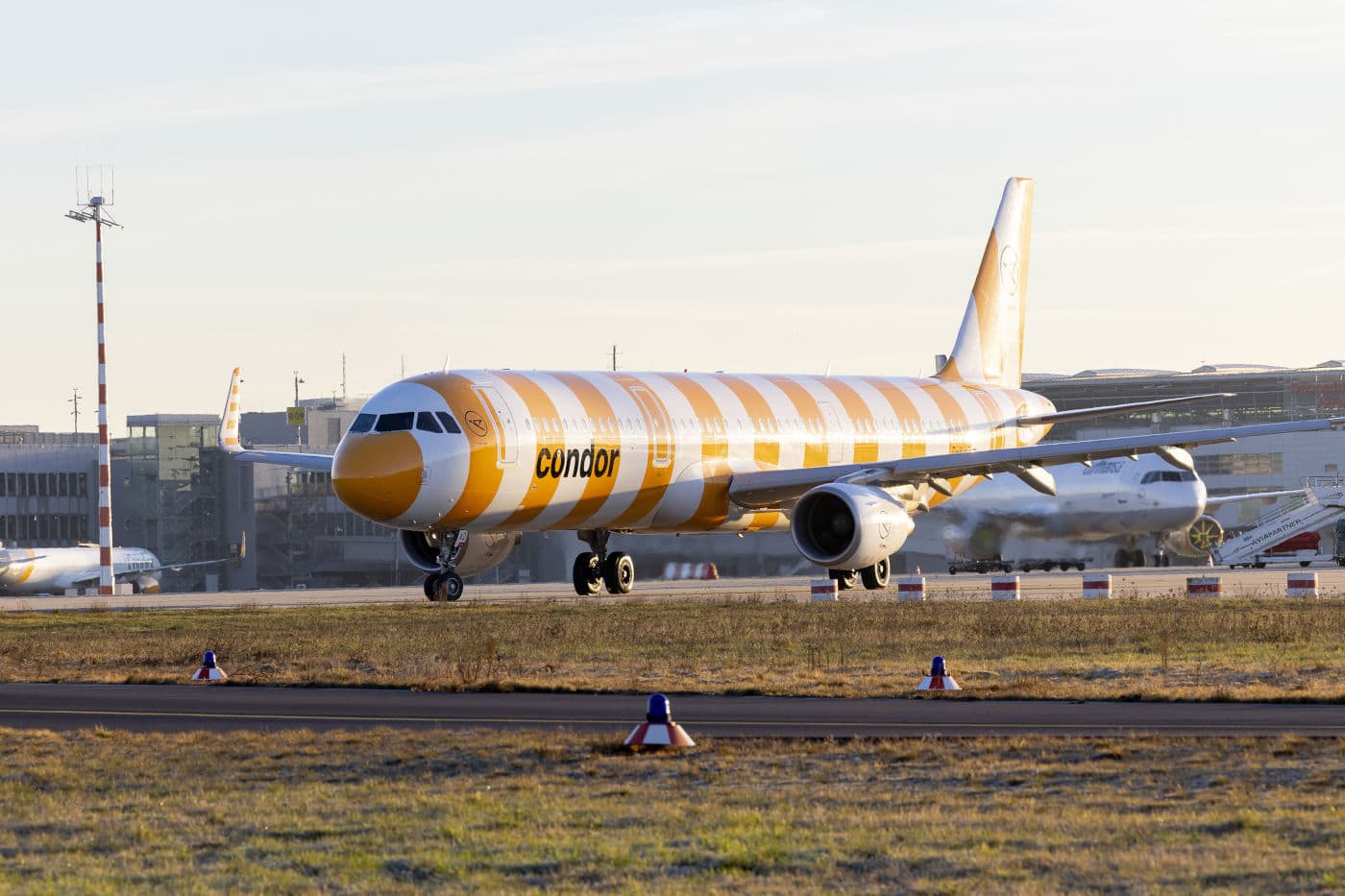 Ein Condor-Flugzeug mit auffälligem gelb-weiß gestreiftem Design steht bei Sonnenaufgang auf dem Vorfeld eines Flughafens, umgeben von weiteren Flugzeugen und Flughafeninfrastruktur. Die Aufnahme zeigt das moderne Passagierflugzeug in voller Länge und betont das neue Lackierungskonzept von Condor Airlines.