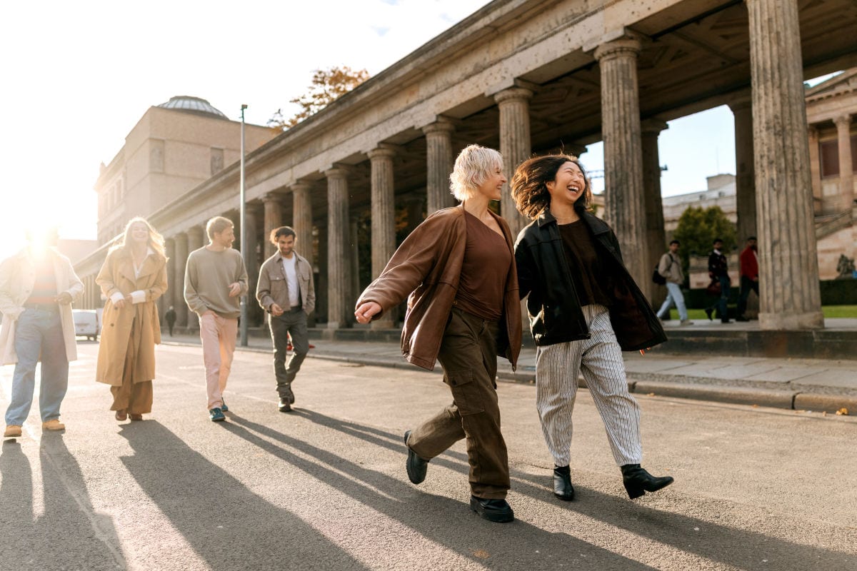Eine Gruppe junger Menschen spaziert gemeinsam bei Sonnenschein vor historischen Säulen in einer europäischen Stadt – ideal für Suchbegriffe wie Gruppenreise, Städtereise, Sightseeing und Freundesgruppe unterwegs.