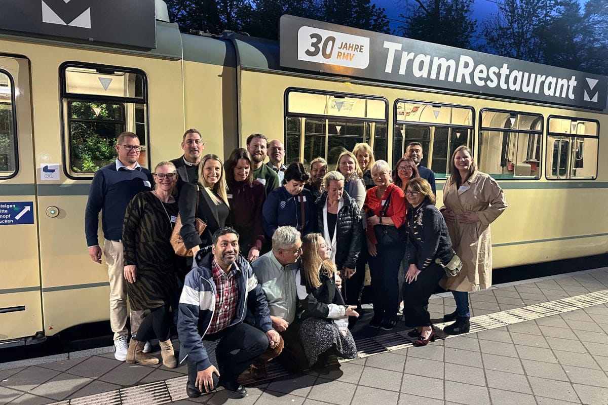 Eine Gruppe von etwa 18 Erwachsenen posiert abends gemeinsam vor einer historischen Straßenbahn mit der Aufschrift „Tram Restaurant“. Die Menschen stehen und knien dicht beieinander auf einem Bahnsteig, lächeln in die Kamera und wirken festlich-schick gekleidet. Die Straßenbahn ist beleuchtet, im Hintergrund sind Bäume im Dämmerlicht zu sehen.