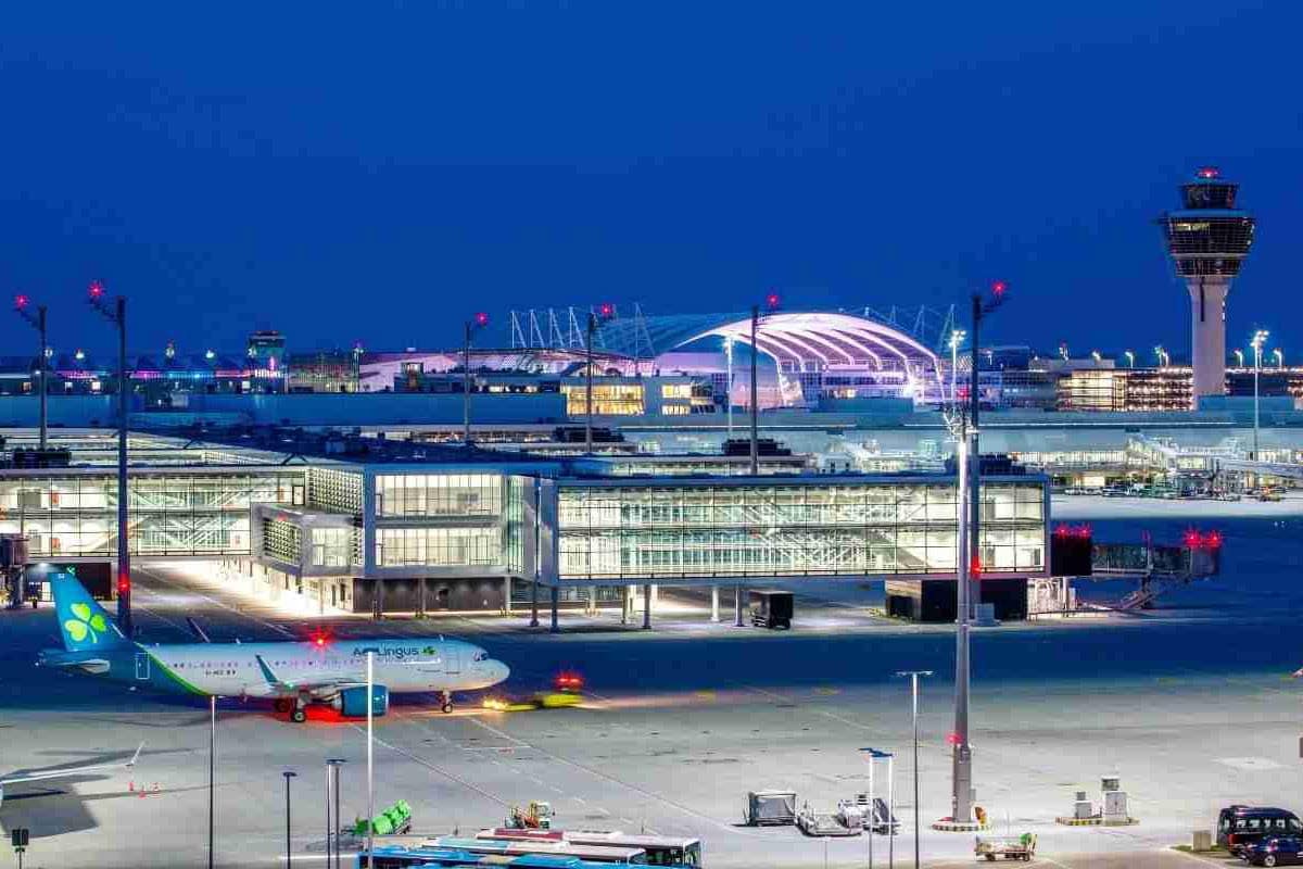 Das Bild zeigt das moderne Terminal 1 des Flughafens München bei Nacht, beleuchtet mit zahlreichen Lichtern und einem zentralen Kontrollturm. Im Vordergrund steht ein Flugzeug von Aer Lingus auf dem Rollfeld.