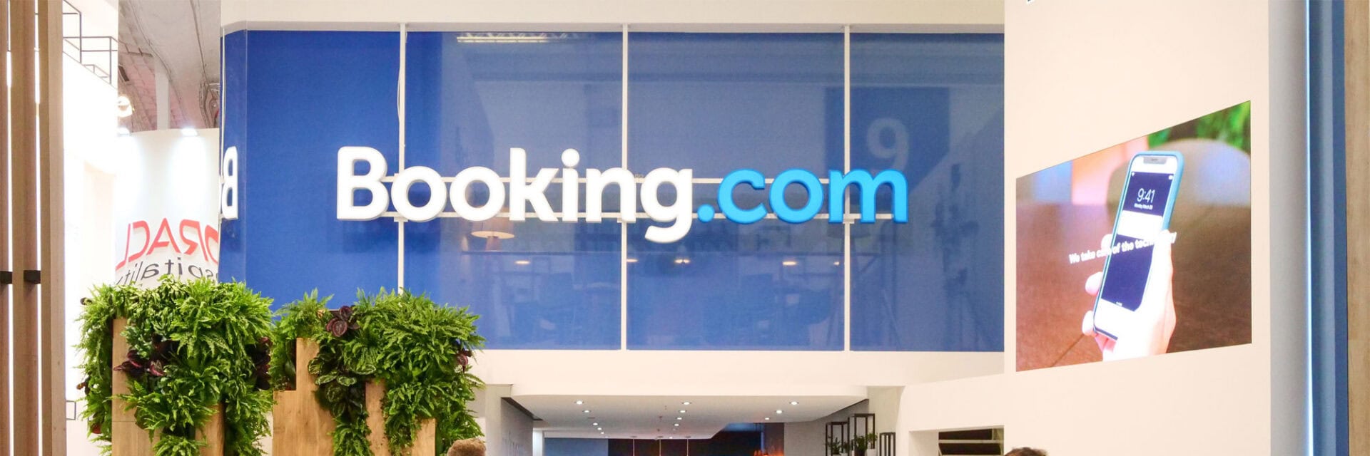 Das Bild zeigt den Messestand von Booking.com auf der ITB 2019 in Berlin, an dem mehrere Besucher und Geschäftspersonen vorbeigehen und sich informieren. Im Vordergrund sind grüne Pflanzen und moderne Einrichtung zu sehen, während das große blaue Booking.com-Logo im Hintergrund hervorsticht. Die Szene vermittelt Messe-Atmosphäre, Networking und die Themen Digitalisierung und Online-Buchung im Tourismus.