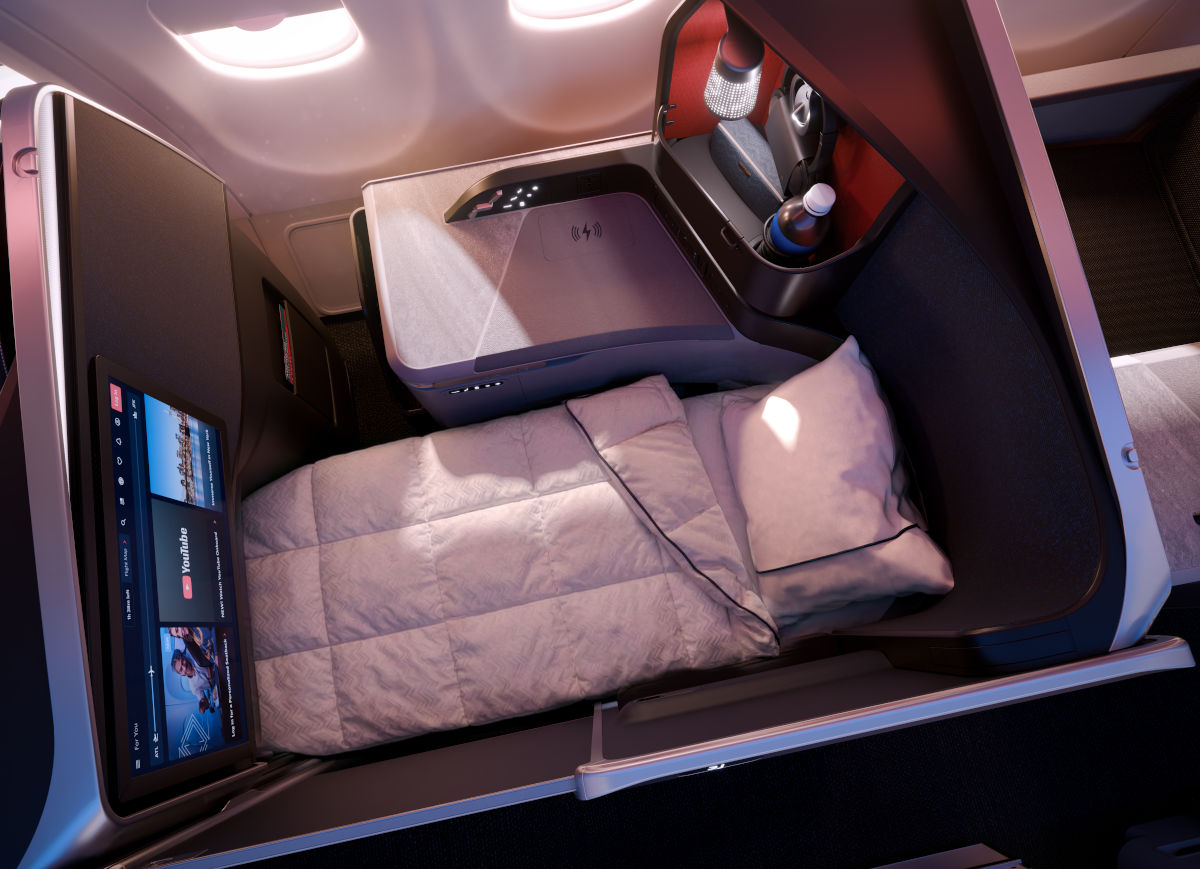 Das Bild zeigt eine luxuriöse Delta One Suite in einem Flugzeug von Delta Air Lines, ausgestattet mit einem flachen Bett, weißen Bettdecken, einem großen Bildschirm und einem modernen Ablagefach. Die Business Class Suite bietet Privatsphäre, Komfort und High-End-Ausstattung für Langstreckenflüge.