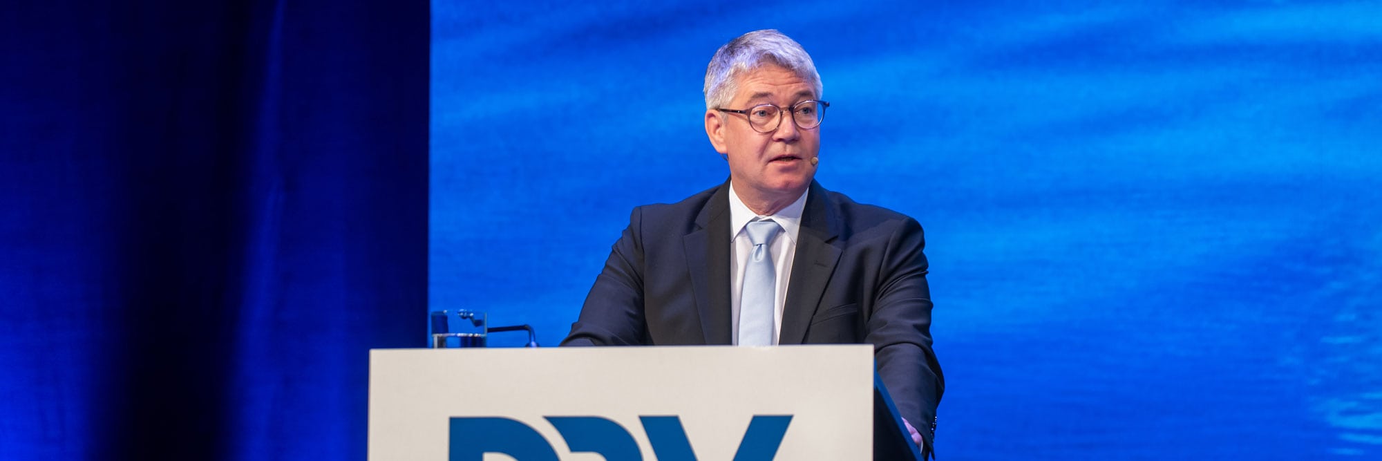 Ein Mann im Anzug steht an einem Rednerpult mit dem Schriftzug „DRV“ und spricht bei einer Konferenz vor blauem Hintergrund. Das Bild zeigt eine typische Szene von einem Kongress oder einer Tagung, bei der ein Redner einen Vortrag hält.