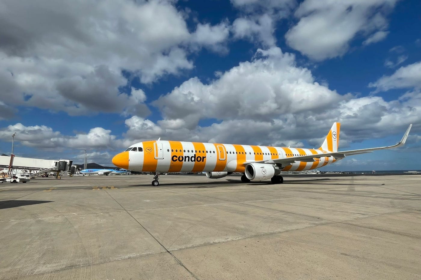 Ein modernes Condor-Flugzeug mit auffälligem gelb-weiß gestreiften Design steht auf dem Rollfeld eines Flughafens unter blauem Himmel mit vereinzelten Wolken. Das Flugzeugmodell ist ein Airbus, bereit für den nächsten Abflug.